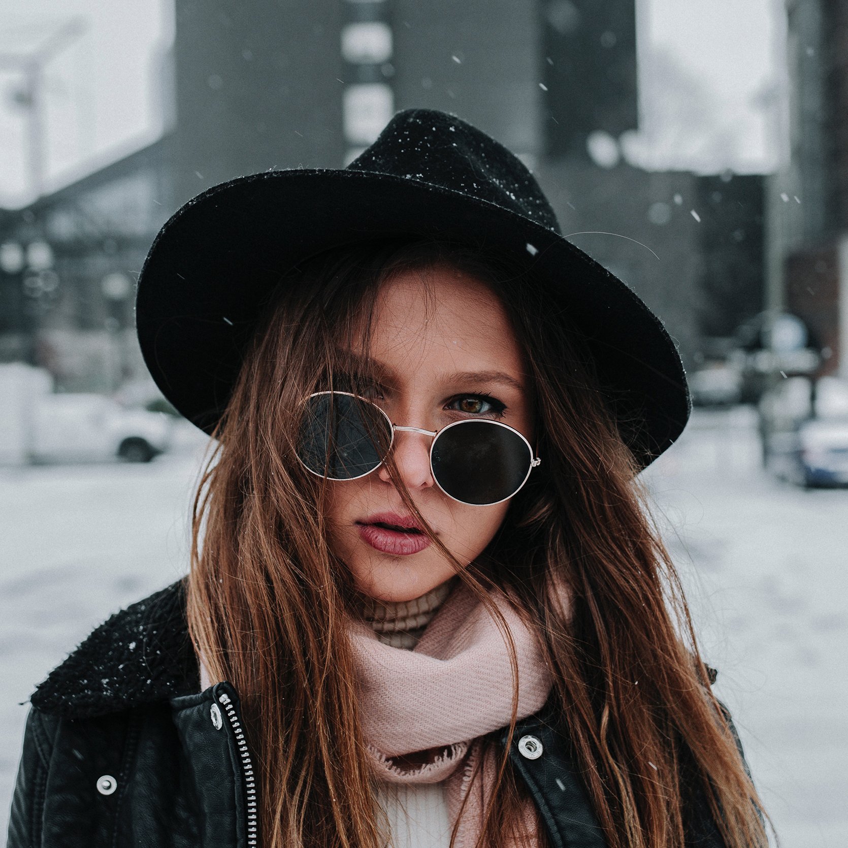 Woman with sunglasses and a black hat in the snow, wearing a jacket and scarf, city background.
